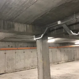 Surowy betonowy strop z zamontowanymi korytami kablowymi i pomarańczową rurą. Widok na instalacje w garażu podziemnym.