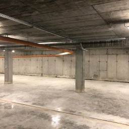 Wszystko w czasie - Surowy betonowy garaż podziemny z pomarańczowymi rurami pod sufitem i metalowymi korytkami kablowymi. Widoczne słupy nośne i betonowa posadzka z śladami.