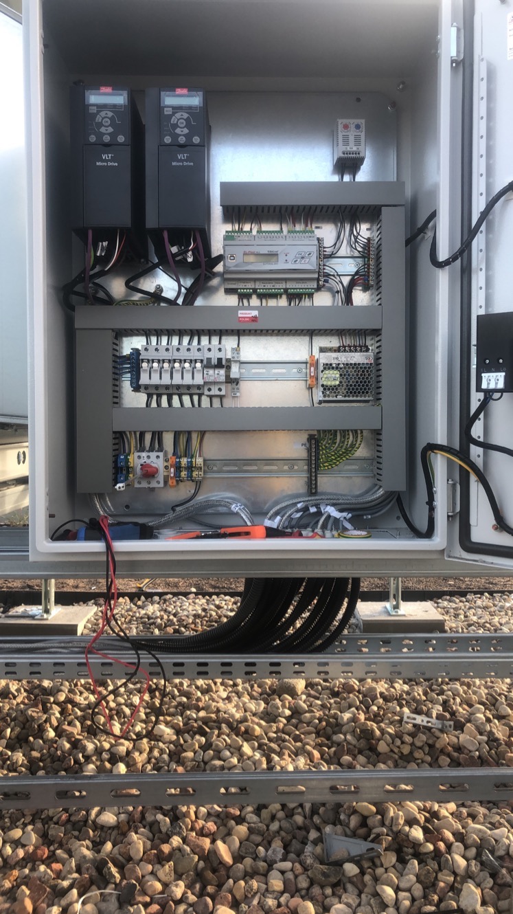 Otwarta szafa sterownicza z widocznymi elementami instalacji elektrycznej, takimi jak sterowniki VLT, bezpieczniki i okablowanie. Widoczne narzędzia w dolnej części szafy.