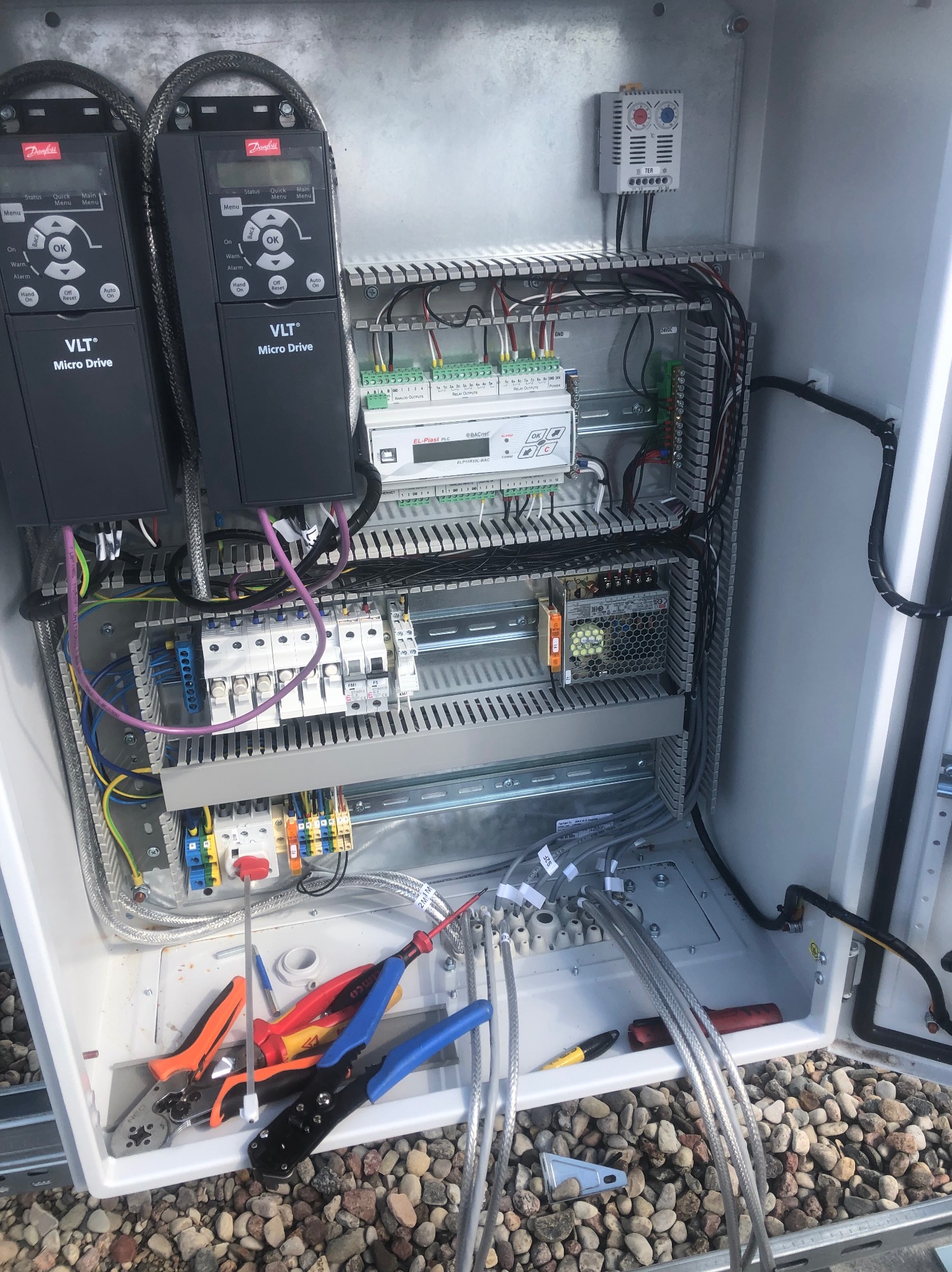 Otwarta szafa sterownicza z dwoma falownikami Danfoss VLT Micro Drive, widoczne kable, bezpieczniki i narzędzia elektryczne. Elementy sterowania wewnątrz obudowy.