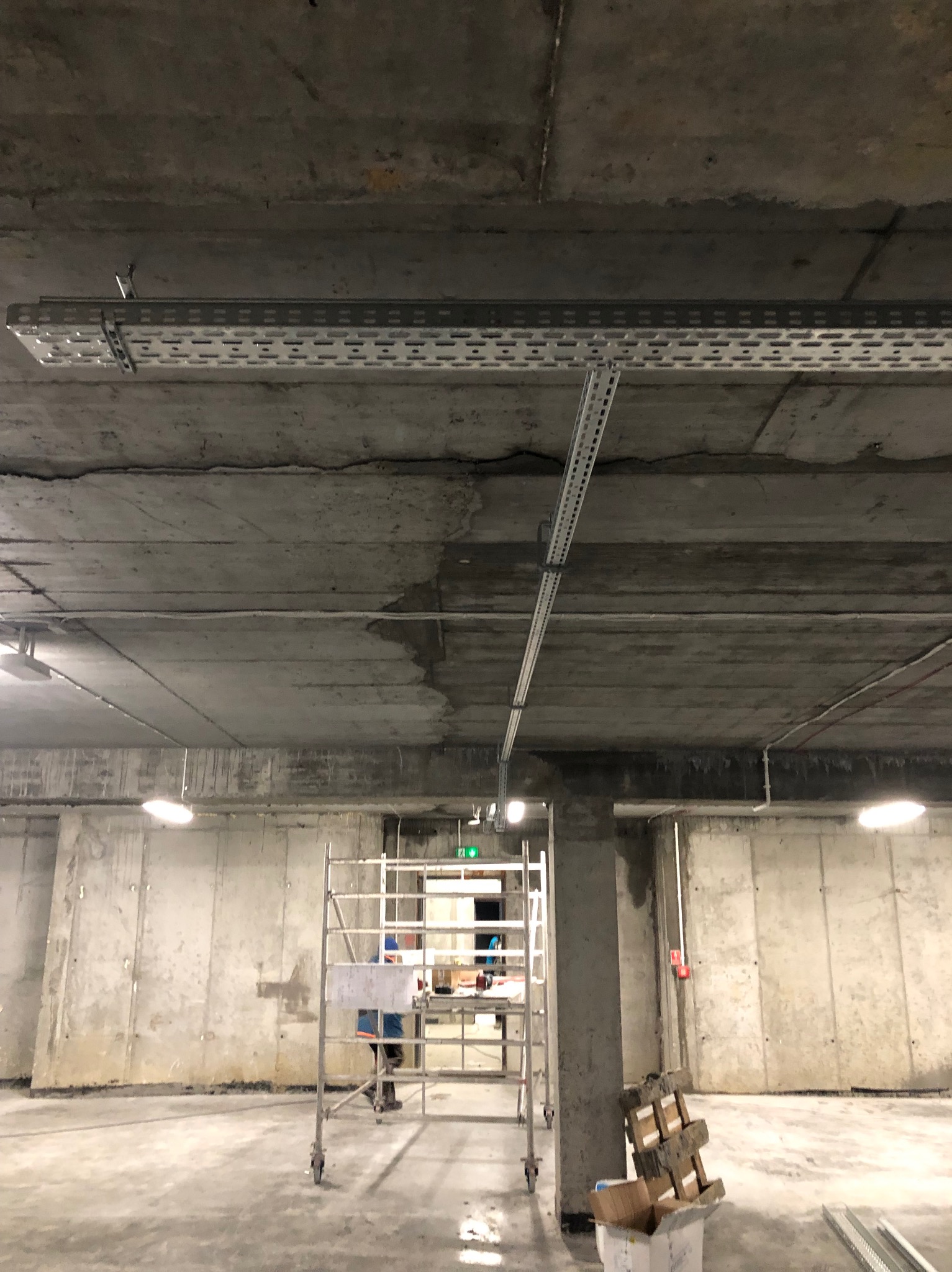 Betonowy strop z zamontowanymi korytkami kablowymi. Widoczne rusztowanie i fragment pracującej osoby. Hala w stanie surowym, oświetlenie sufitowe.