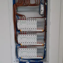 Rozdzielnica elektryczna w białej obudowie, widoczne bezpieczniki, przewody w kolorach brązowym, niebieskim, żółto-zielonym, zamontowana na białej ścianie.