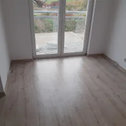 Pusty pokój z panelami podłogowymi w odcieniach beżu, białe ściany i dwuskrzydłowe okno balkonowe z widokiem na teren budowy.