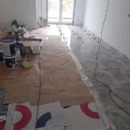 Wnętrze w trakcie układania szarych płytek podłogowych z użyciem poziomujących klipsów, widoczne narzędzia i materiały budowlane zabezpieczone kartonem.