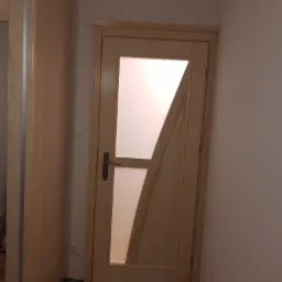 Drewniane drzwi wewnętrzne z asymetrycznym przeszkleniem, zamontowane w jasnym pomieszczeniu, widoczne zabezpieczenia podłogi podczas prac wykończeniowych.