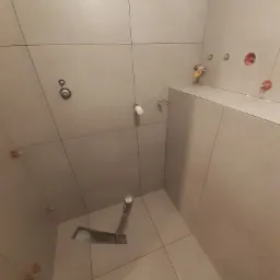 Świeżo ułożone, duże, beżowe płytki w małej łazience z widocznymi elementami instalacji wodnej i kanalizacyjnej oraz plastikowymi krzyżykami dystansowymi.