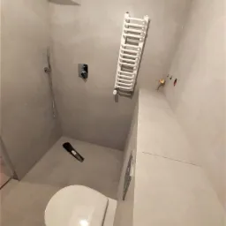 Nowoczesna łazienka z kabiną prysznicową typu deszczownica, grzejnikiem drabinkowym i podtynkową spłuczką WC, wykończona płytkami w odcieniach szarości, z widocznymi otworami instalacyjnymi.