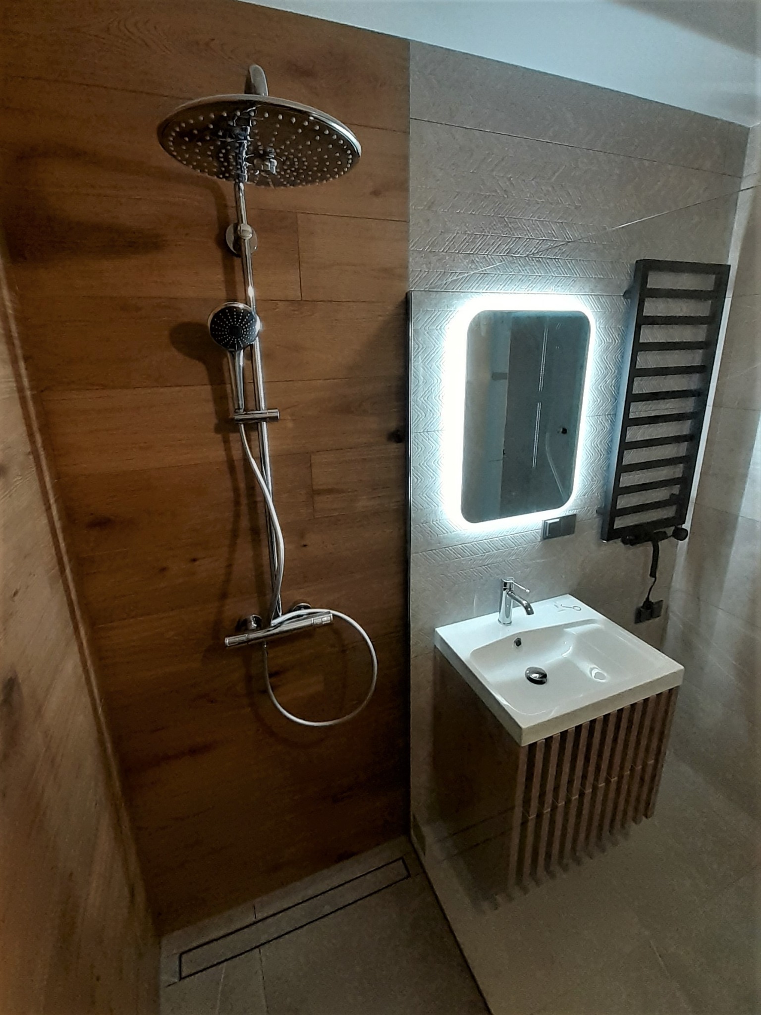 Nowoczesna łazienka z drewnianą ścianą prysznicową, deszczownicą, umywalką z szafką z drewnianych listewek i podświetlanym lustrem.