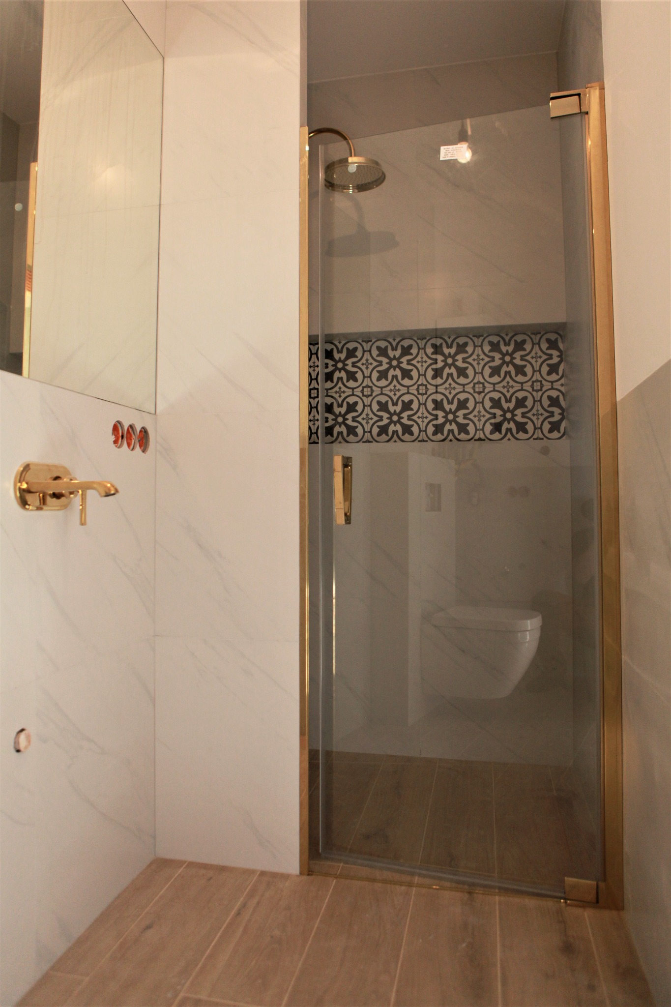 Nowoczesna łazienka z kabiną prysznicową z złotymi profilami, widoczna bateria umywalkowa w kolorze złota, płytki imitujące marmur oraz dekoracyjny pas płytek nad toaletą.