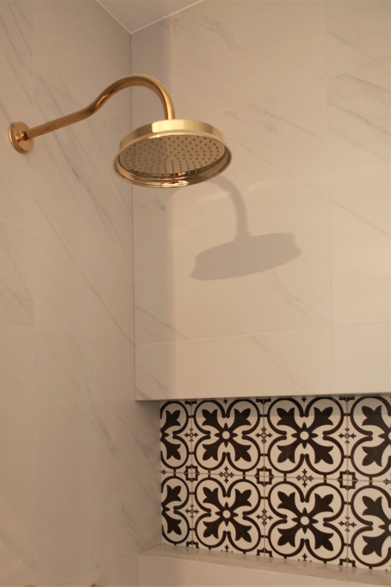 Złota deszczownica zamontowana na ścianie wyłożonej marmurowymi płytkami, z dekoracyjną wnęką wykończoną czarno-białymi płytkami w geometryczne wzory.