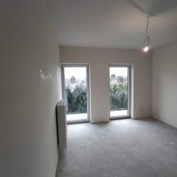 IMK GROUP Sp. z o.o. - Pokój w trakcie remontu: ściany po szpachlowaniu, surowa podłoga, dwa okna z widokiem na zieleń, grzejnik przy ścianie, wisząca żarówka bez oprawy.