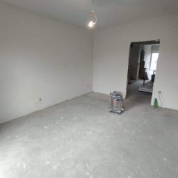 IMK GROUP Sp. z o.o. - Pokój w trakcie przygotowania do malowania: wyszpachlowane ściany, surowa podłoga, widoczny odkurzacz przemysłowy i prowizoryczne oświetlenie.