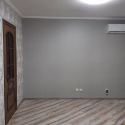 IMK GROUP Sp. z o.o. - Pomalowane na szaro wnętrze pokoju z panelami podłogowymi imitującymi drewno, brązowe drzwi z szybą, klimatyzacja na ścianie i okrągła lampa sufitowa.