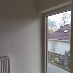 IMK GROUP Sp. z o.o. - Ściana po szpachlowaniu, widoczny grzejnik, okno PCV z widokiem na dom z brązową dachówką i drzewo bez liści.