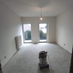 IMK GROUP Sp. z o.o. - Pokój w trakcie przygotowania do malowania, z widocznym odkurzaczem przemysłowym pośrodku i surowymi ścianami, z dwoma oknami wychodzącymi na zieleń