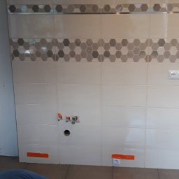 IMK GROUP Sp. z o.o. - Ściana w trakcie układania płytek w kuchni, widoczne otwory na instalacje, dekoracyjny pas z heksagonami, pomarańczowa taśma zabezpieczająca na płytkach i gniazdkach.