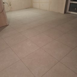 IMK GROUP Sp. z o.o. - Wyłożona jasnymi, kwadratowymi płytkami podłoga w surowym pomieszczeniu z widocznymi instalacjami elektrycznymi i oknem.