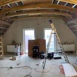 IMK GROUP Sp. z o.o. - Poddasze w trakcie ocieplania wełną mineralną, widoczne drewniane krokwie, metalowe profile, dwa okrągłe okna, drabina, oświetlenie robocze i narzędzia.