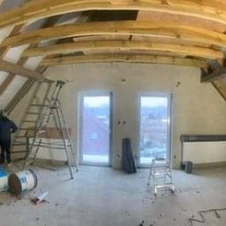 IMK GROUP Sp. z o.o. - Poddasze w trakcie adaptacji: ocieplenie skośnego dachu wełną mineralną, drewniane belki konstrukcyjne, okrągłe okno, drabina, narzędzia i materiały budowlane rozrzucone na podłodze.