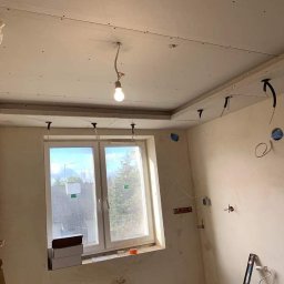 IMK GROUP Sp. z o.o. - Wnętrze w trakcie remontu: sufit podwieszany z płyt gipsowo-kartonowych w trakcie montażu, widoczne kable, okno z widokiem na drzewa, drabina oparta o ścianę, podłoga z białymi płytkami.