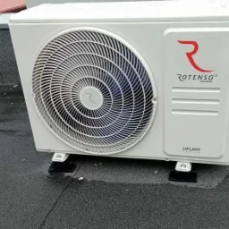 Biała jednostka zewnętrzna klimatyzacji Rotenso UKURA na dachu pokrytym papą, zamontowana na czarnych wspornikach antywibracyjnych. Widoczne ślady deszczu.