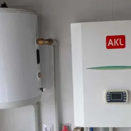 Biały, wiszący bojler i piec gazowy AKL na tle białych płytek. Widoczne rury i przewody. Minimalistyczny design.