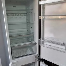 ST-TRONIC Łukasz Stojak - Pusta, biała lodówka Miele z otwartymi drzwiami, ukazująca półki, szuflady i uchwyt na butelki. Widoczny panel sterowania na górze.