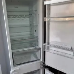 Pusta, biała lodówka Miele z otwartymi drzwiami, ukazująca półki, szuflady i uchwyt na butelki. Widoczny panel sterowania na górze.