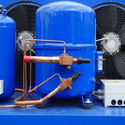 ST-TRONIC Łukasz Stojak - Niebieski agregat chłodniczy z miedzianymi rurami i wentylatorami w tle. Widoczne elementy sterujące i skrzynka elektryczna. Sprzęt chłodniczy w zbliżeniu.