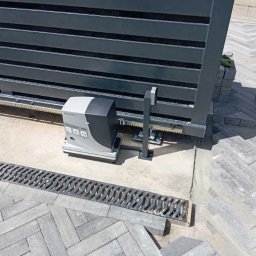 ST-TRONIC Łukasz Stojak - Szara, przesuwna brama z automatyką, widoczny mechanizm napędowy i listwa zębata. Kostka brukowa ułożona w jodełkę, odwodnienie liniowe. Brama w trakcie montażu.