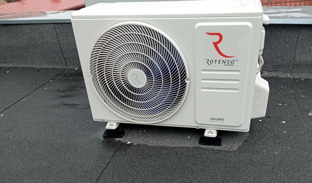 Biała jednostka zewnętrzna klimatyzacji Rotenso UKURA na dachu pokrytym papą, zamontowana na czarnych wspornikach antywibracyjnych. Widoczne ślady deszczu.