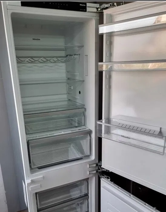 Pusta, biała lodówka Miele z otwartymi drzwiami, ukazująca półki, szuflady i uchwyt na butelki. Widoczny panel sterowania na górze.
