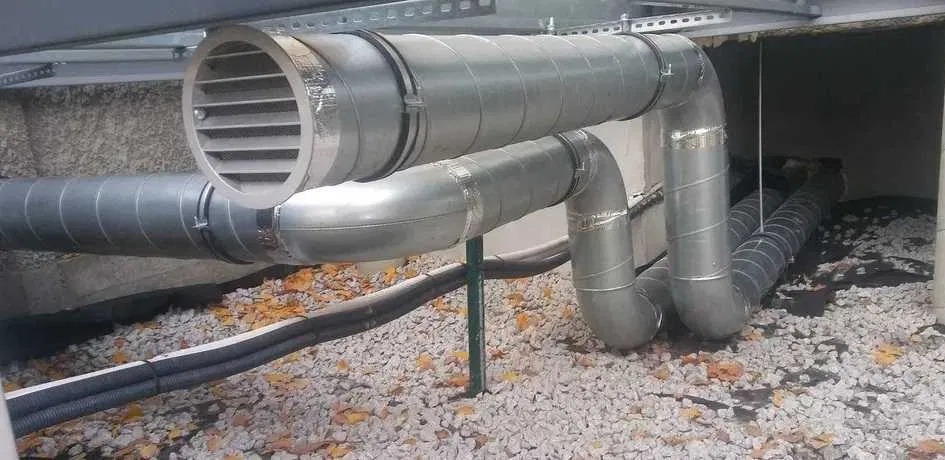 Szare, metalowe rury wentylacyjne z okrągłym wylotem, zamontowane pod sufitem, z widocznymi elementami mocującymi i izolacją, na tle szarego żwiru i opadłych liści.