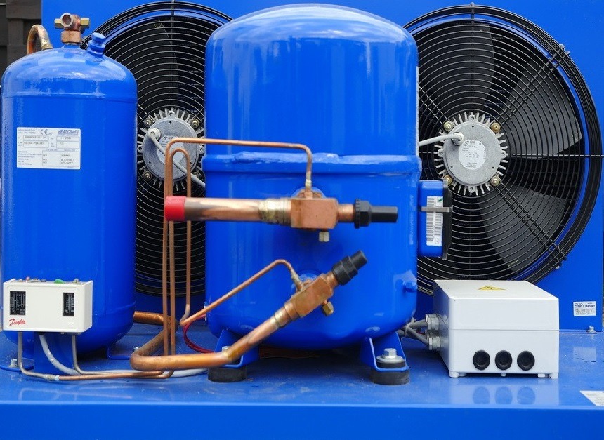 Niebieski agregat chłodniczy z miedzianymi rurami i wentylatorami w tle. Widoczne elementy sterujące i skrzynka elektryczna. Sprzęt chłodniczy w zbliżeniu.