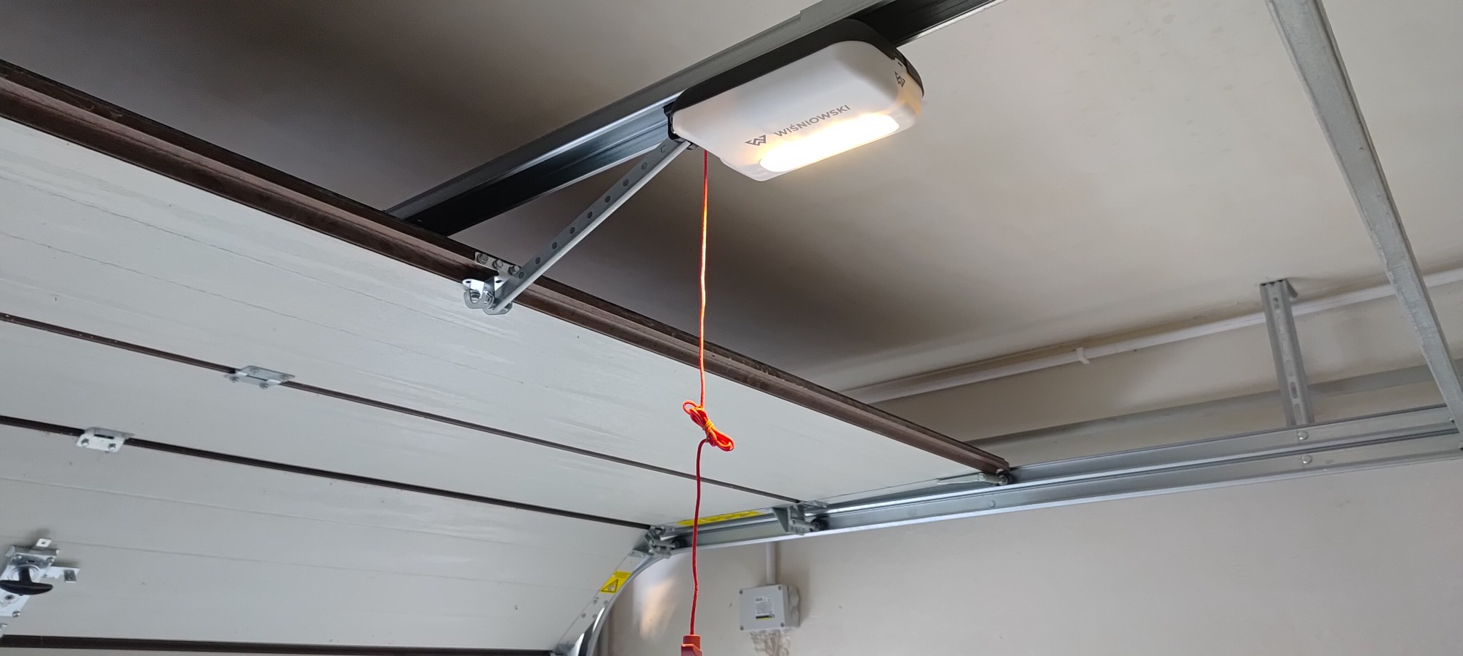 Zamknięta brama garażowa Wiśniowski z widocznym napędem i oświetleniem, zamontowana pod sufitem garażu. Widok od wewnątrz, z perspektywy osoby stojącej w garażu.