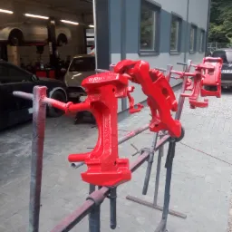 Czerwone, świeżo pomalowane zaciski hamulcowe na metalowym stojaku przed warsztatem samochodowym, w tle podnośnik z autem bez kół i zaparkowany czarny samochód.