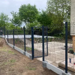 Nowo postawione, metalowe ogrodzenie panelowe z bramką, osadzone na betonowej podmurówce. W tle widoczne drzewa i częściowo zagospodarowany teren z folią. Na pierwszym planie narzędzia i preparaty...