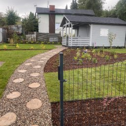 LEWERTE SPÓŁKA Z OGRANICZONĄ ODPOWIEDZIALNOŚCIĄ - Ogród z szarą altaną, ścieżką z kamieni i żwiru oraz rabatami kwiatowymi, ogrodzony czarnym panelem. W tle widoczny dom z anteną.