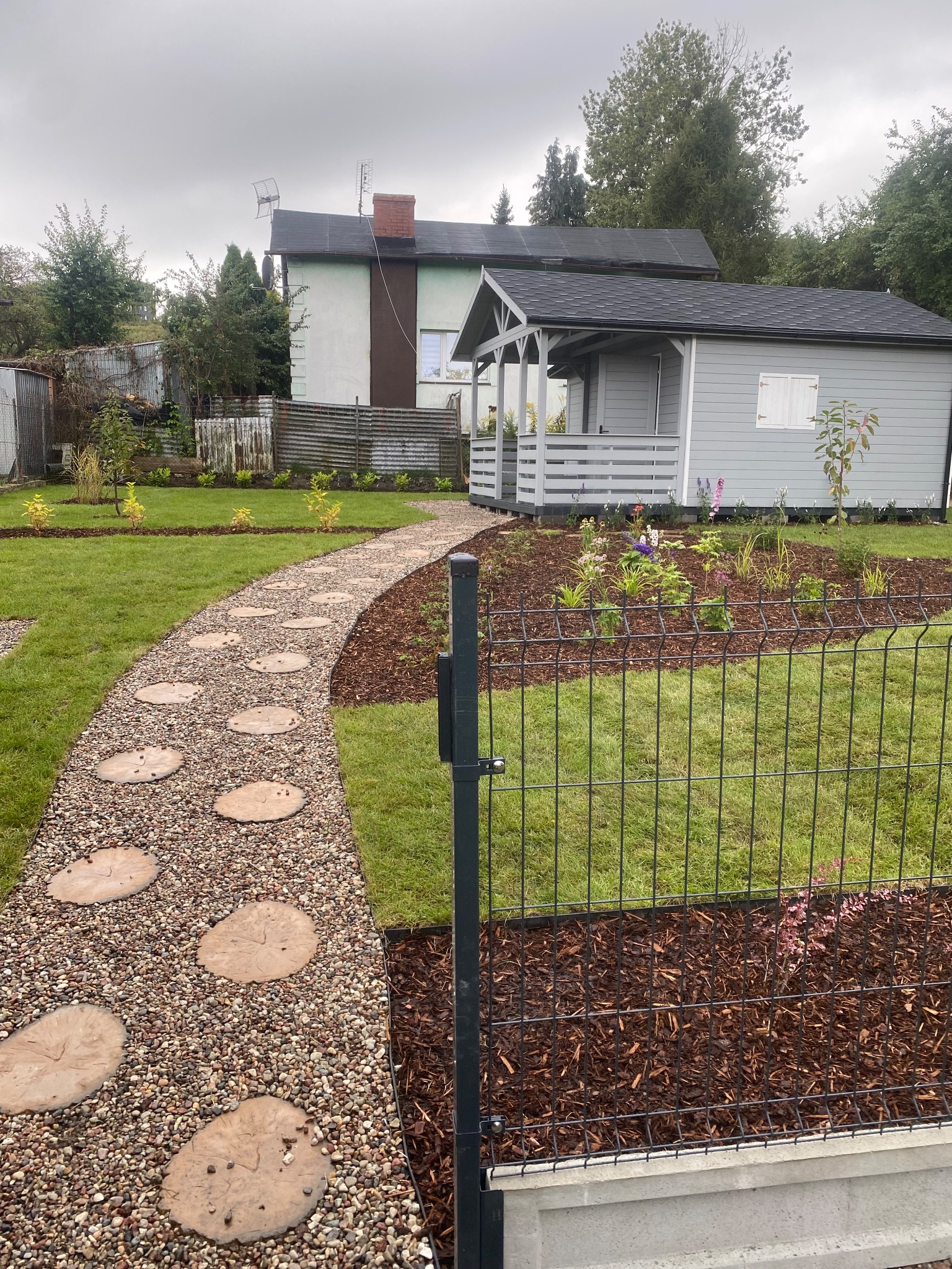 Ogród z szarą altaną, ścieżką z kamieni i żwiru oraz rabatami kwiatowymi, ogrodzony czarnym panelem. W tle widoczny dom z anteną.
