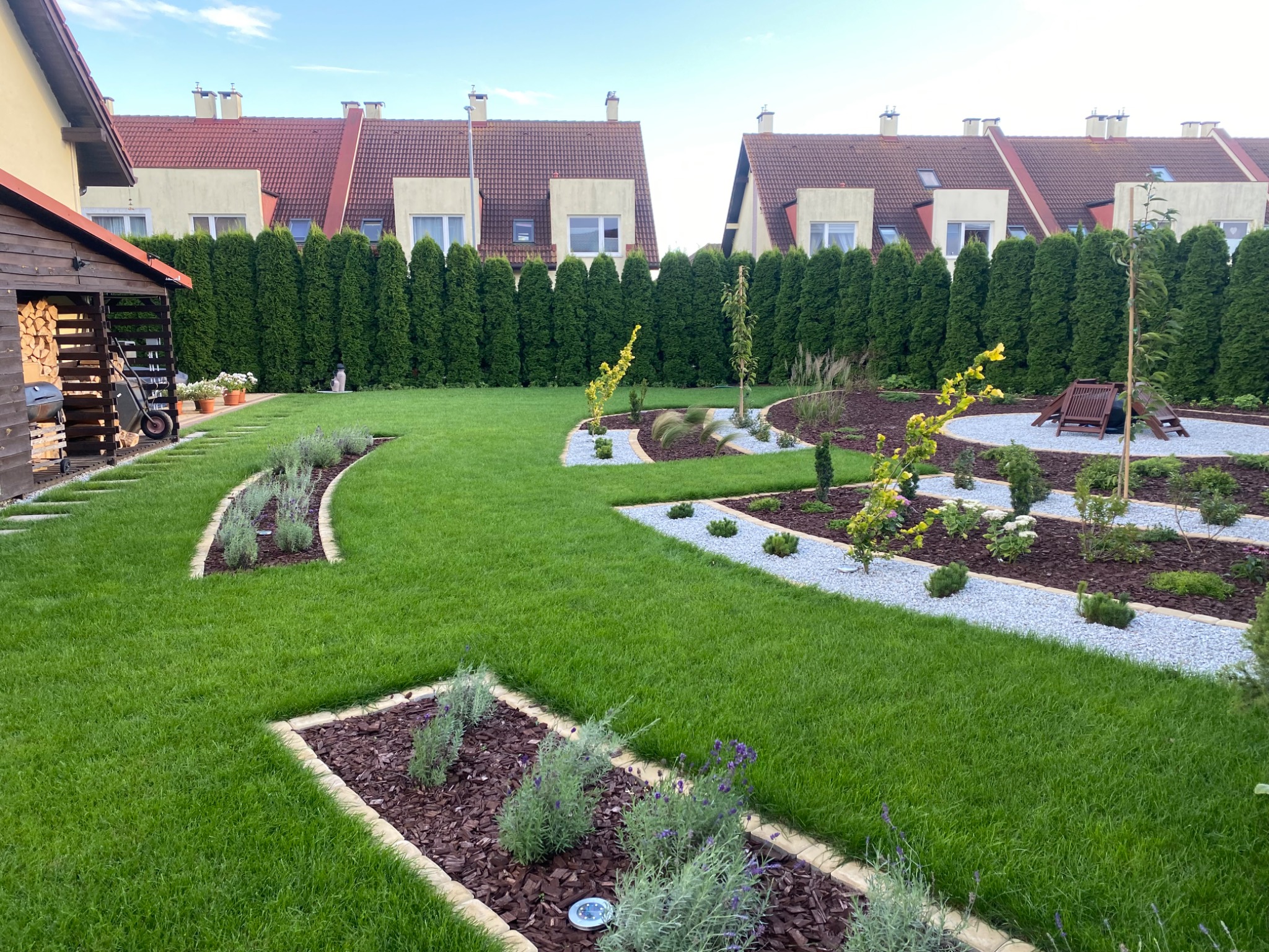 Zadbany ogród z równo przystrzyżonym trawnikiem, ozdobnymi rabatami z kamykami, korą i roślinami. W tle rząd wysokich tuj i zabudowa szeregowa.