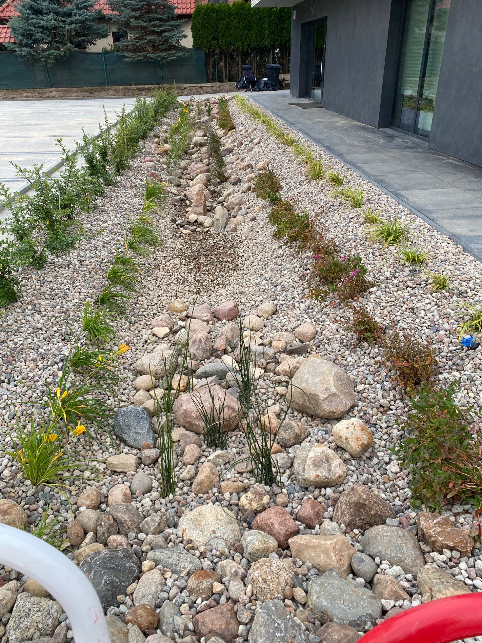 Ogród z kamieniami i roślinami ozdobnymi wzdłuż nowoczesnego domu. Widoczne różne gatunki roślin i kamieni, tworzące estetyczną aranżację.