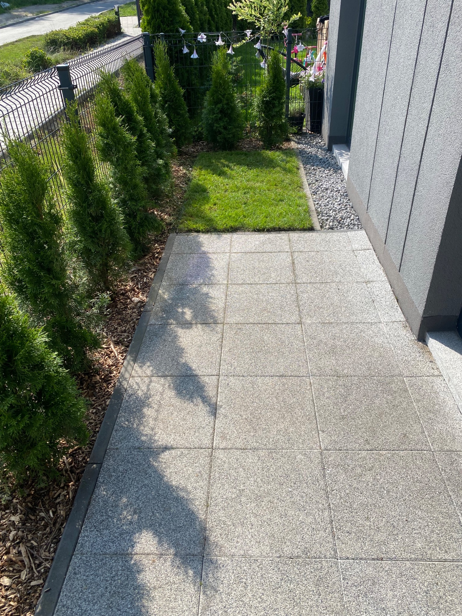 Ułożona kostka granitowa tworząca ścieżkę wzdłuż domu, obok trawnik i rząd tuj. Widoczne elementy dekoracyjne w ogrodzie. Słoneczny dzień.