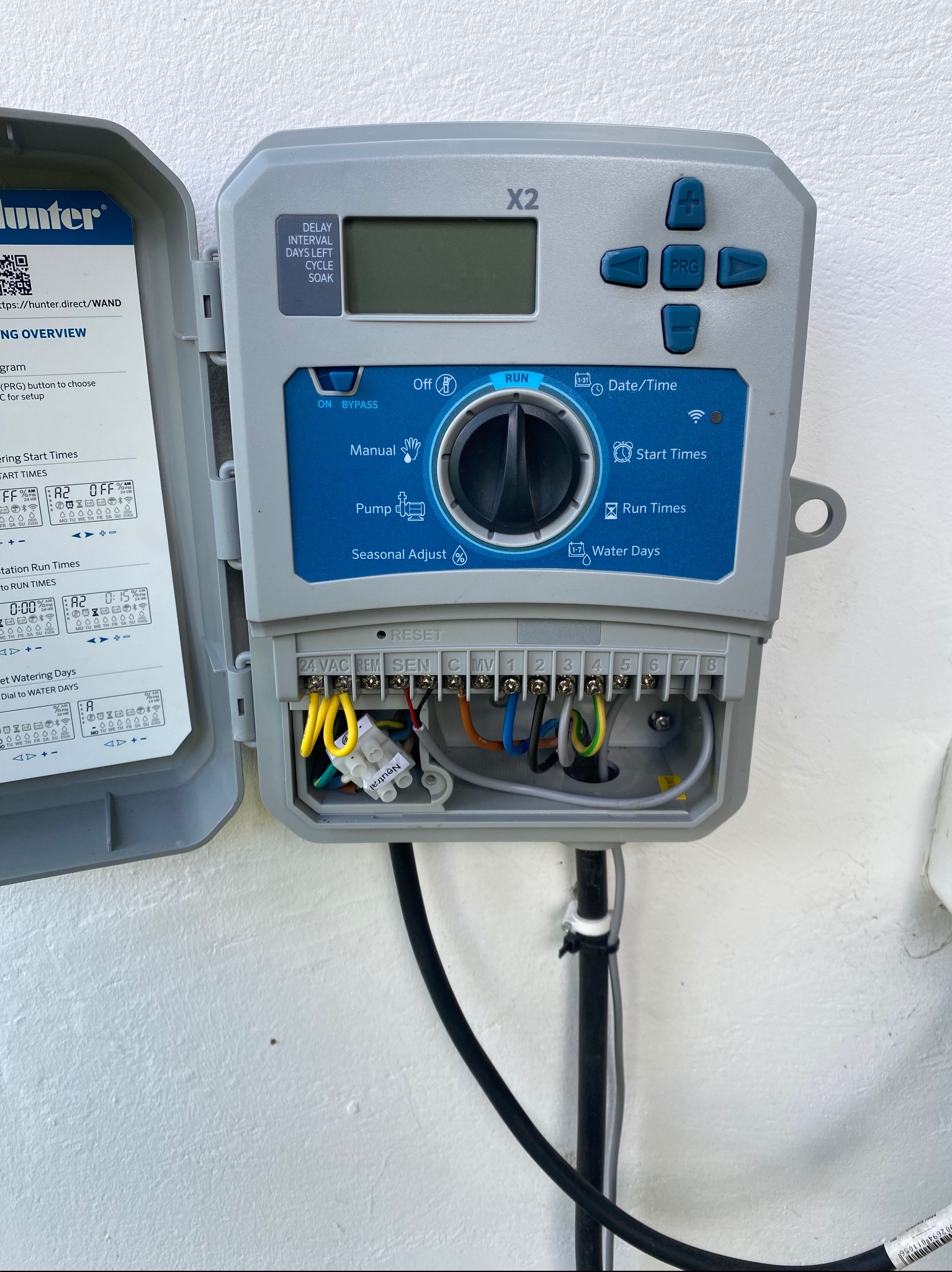 Szary sterownik nawadniania Hunter X2 z otwartą obudową, widoczne okablowanie i instrukcja. Panel sterowania z przyciskami i wyświetlaczem.