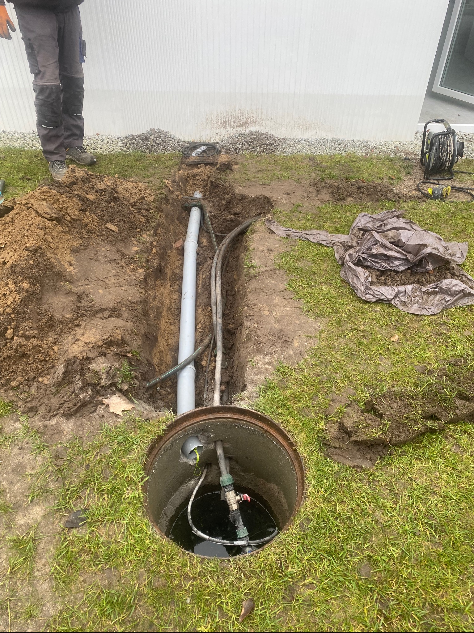 Studzienka rewizyjna z rurami w wykopie na trawniku, widoczne elementy instalacji nawadniającej i kabel elektryczny. Prace ziemne w ogrodzie.