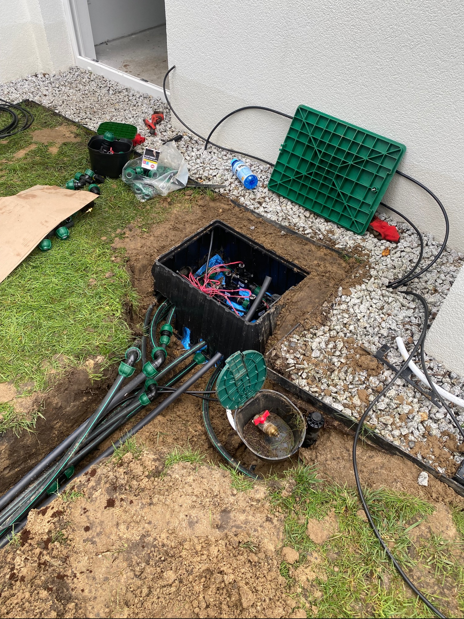 Instalacja systemu nawadniania w Gdańsku: wykopana skrzynka z przewodami, rurami i akcesoriami na trawniku. Widoczne elementy systemu przed zakopaniem.