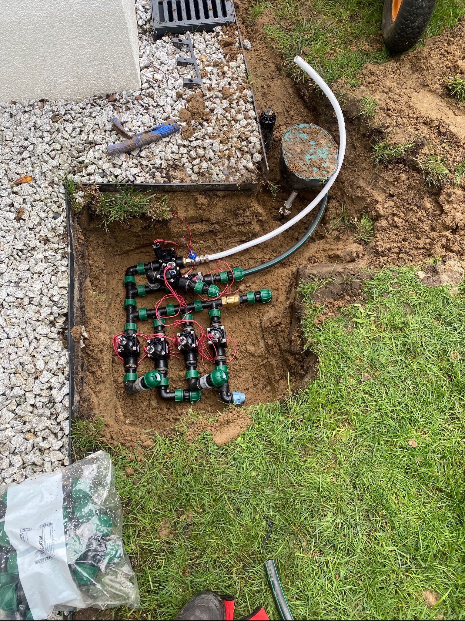 Wyeksponowany system nawadniania w wykopie, z widocznymi rurami, złączkami i przewodami elektrycznymi. Częściowo widoczna trawa i kamienie dekoracyjne.
