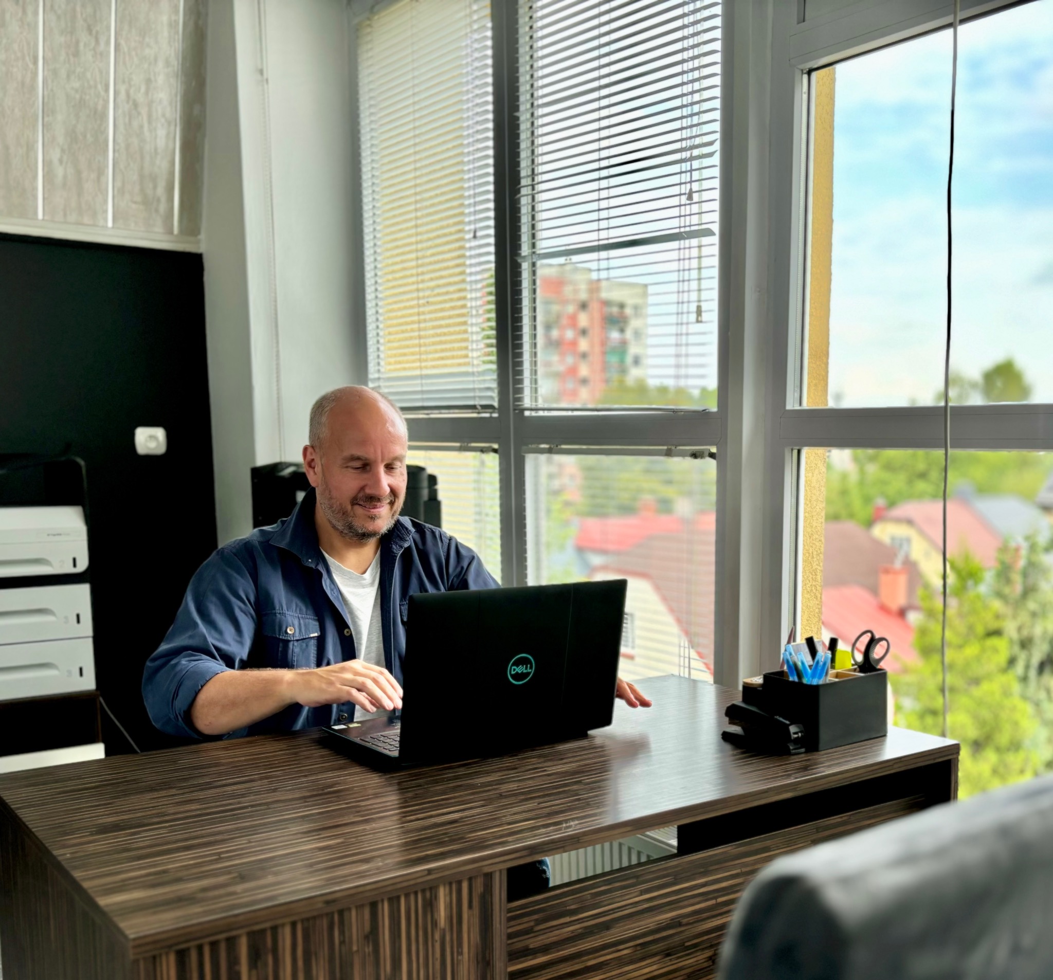 Mężczyzna w niebieskiej koszuli pracuje na laptopie Dell przy biurku z organizerem na długopisy i zszywaczem; za oknem widok na budynek mieszkalny.