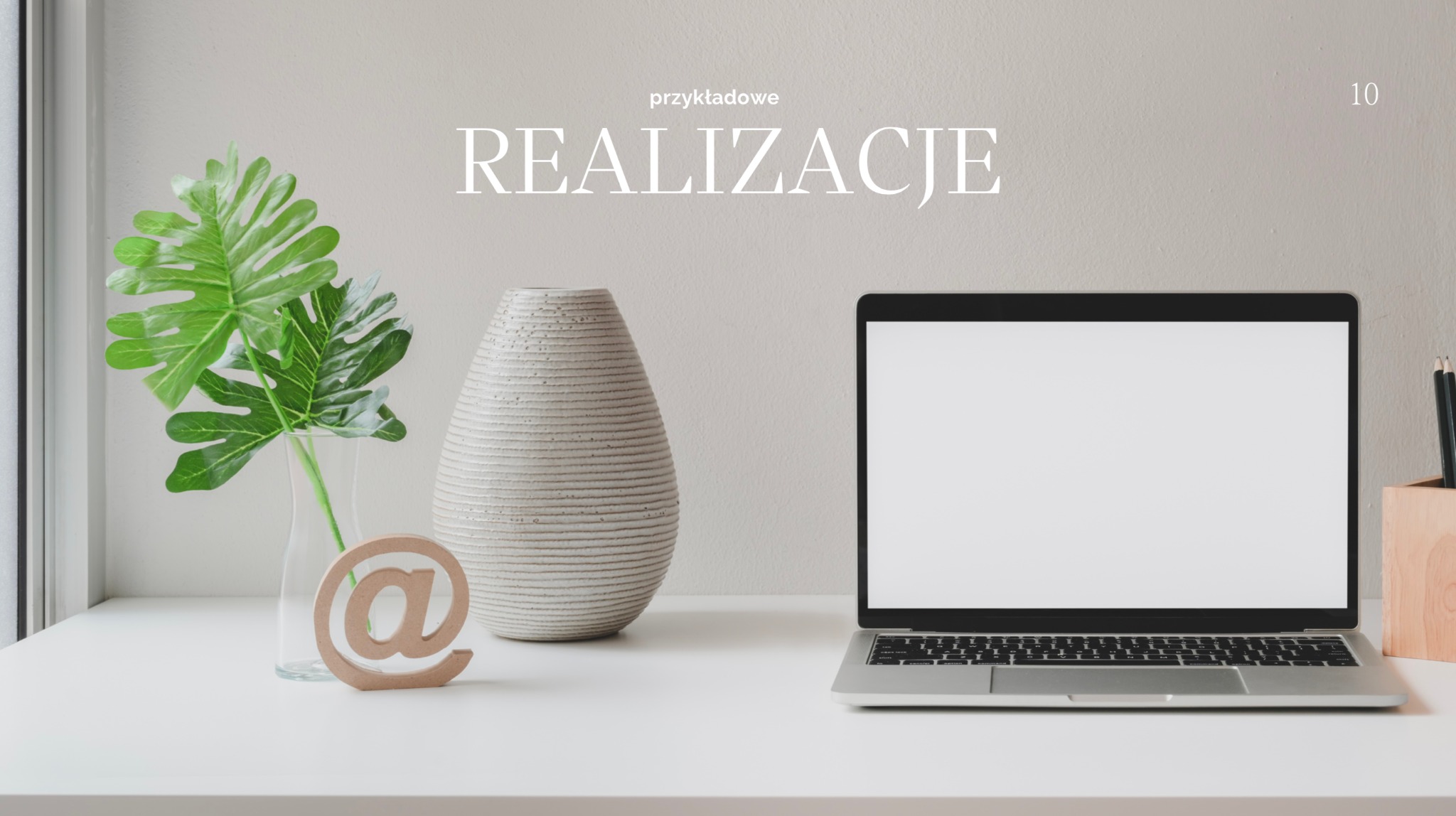 Minimalistyczne biurko z laptopem z pustym ekranem, wazonem z zielonymi liśćmi i drewnianym symbolem @, sugerujące realizacje online.