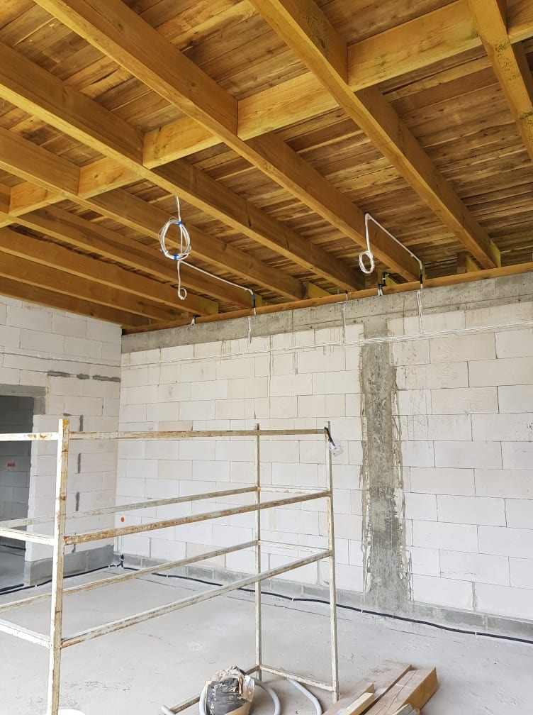 Surowy stan wnętrza z widoczną drewnianą konstrukcją stropu, wyprowadzonymi przewodami elektrycznymi zwisającymi z sufitu i przewodami ułożonymi wzdłuż ściany z pustaków, oraz fragment rusztowania.