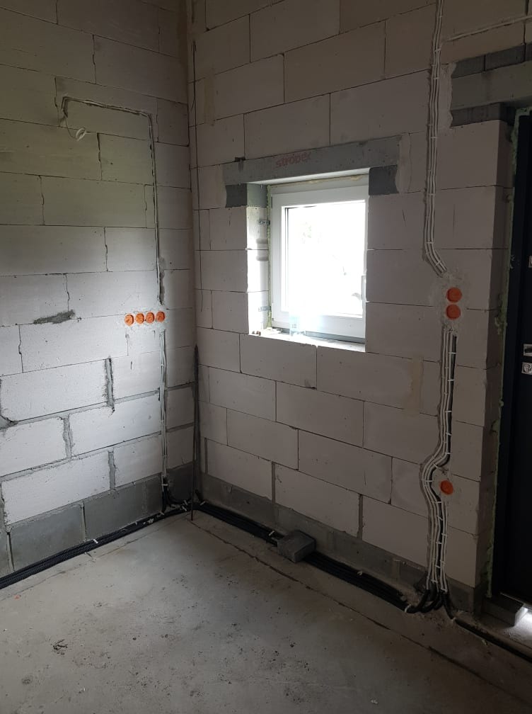 Surowy stan wnętrza z widocznymi instalacjami elektrycznymi: kable w peszlach prowadzone po ścianach z bloczków, puszki elektryczne osadzone w ścianie, okno z parapetem. Widoczne również czarne...